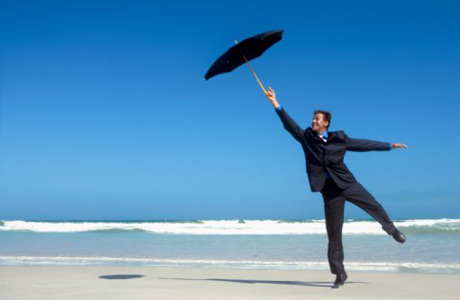 Mann im Anzug am Strand, mit Schirm in der Hand, wird vom Wind verweht.